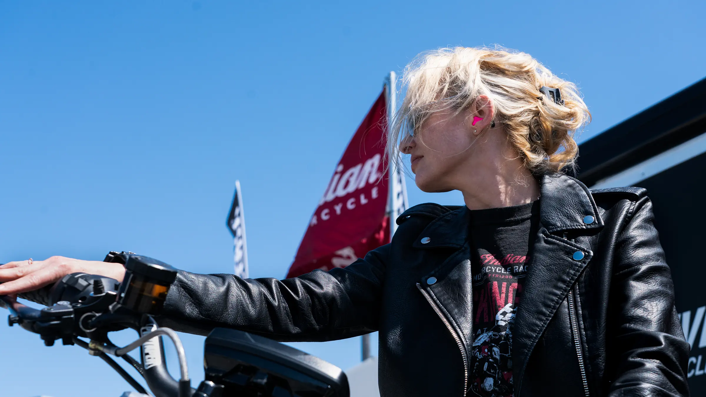 Rider in a leather jacket reaching for her handlebars, a pink Big Ear custom earplug in her ear