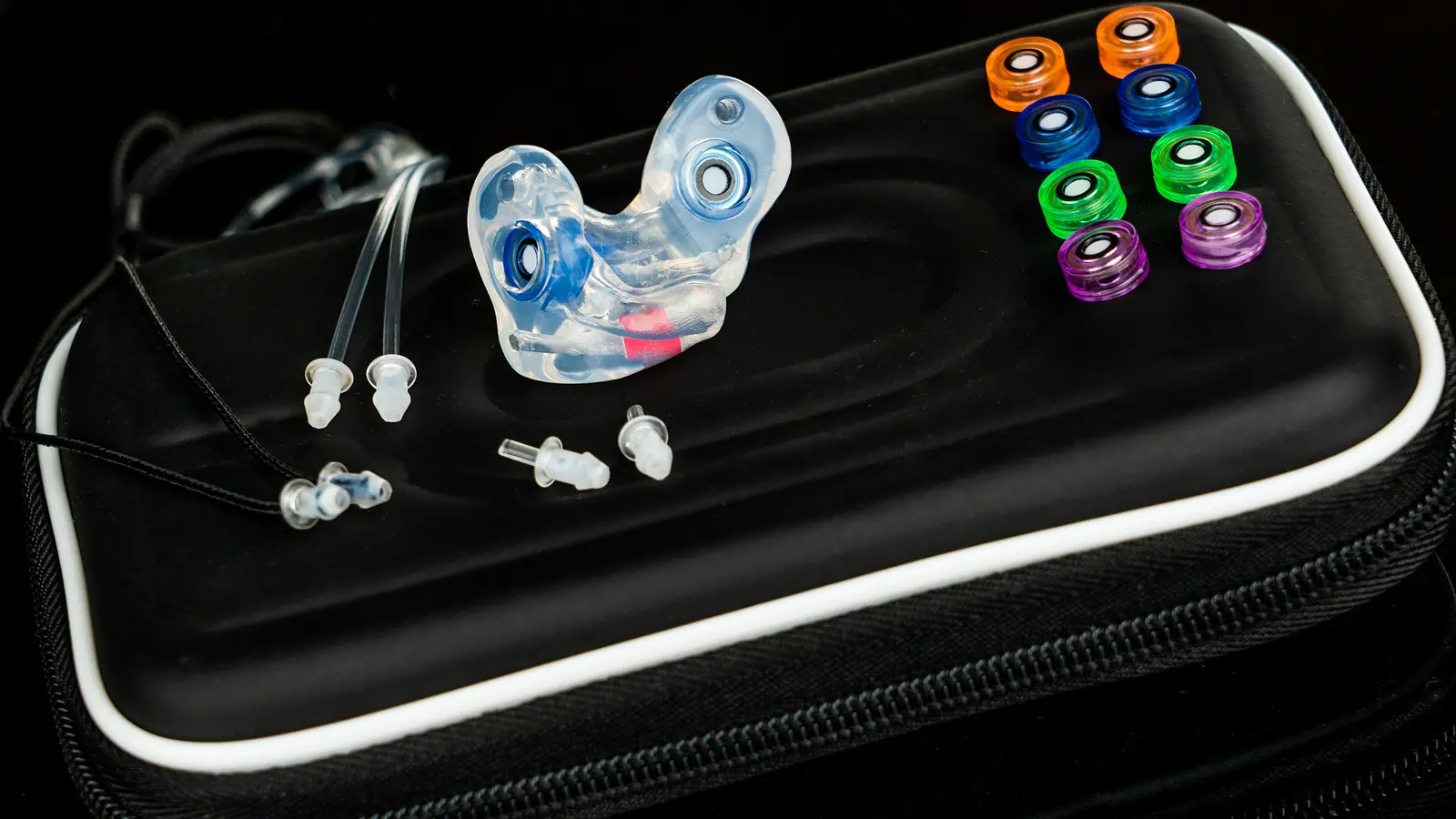 The One custom filtered earplugs in clear silicone on a black travel case beside spare filters in orange, blue, green and purple