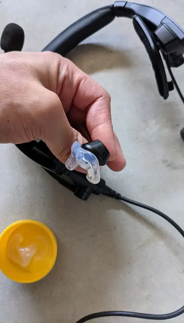 A hand holding the Bose ProFlight earbud seated in its custom adapter, headset and case nearby