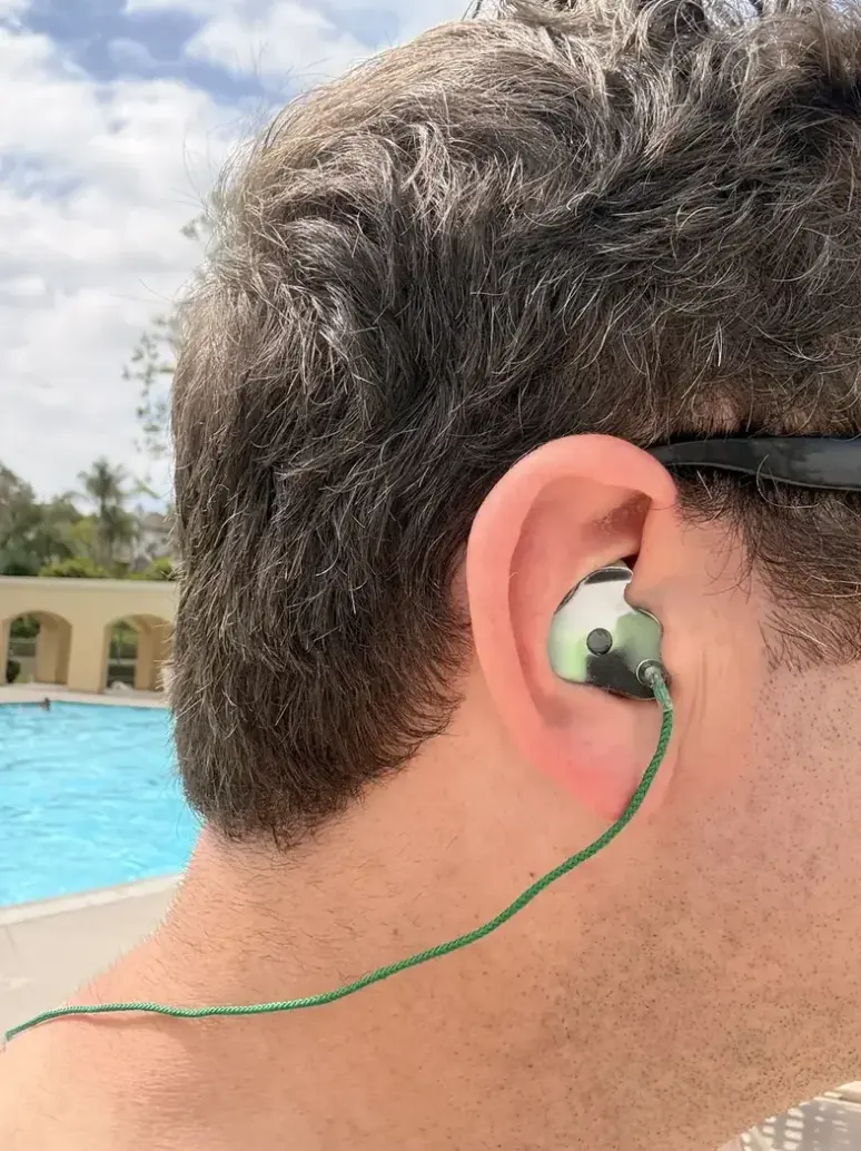 A customer wearing a green camo custom swimming earplug, pool in the background